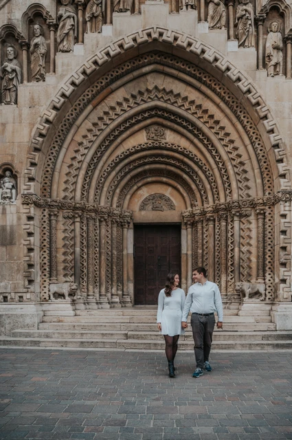 A walk beneath the grand stone arches at a Budapest anniversary photoshoot The couple walking towards the camera with the chapel’s grand arched entrance at Vajdahunyad Castle behind them during their anniversary photoshoot in Budapest.