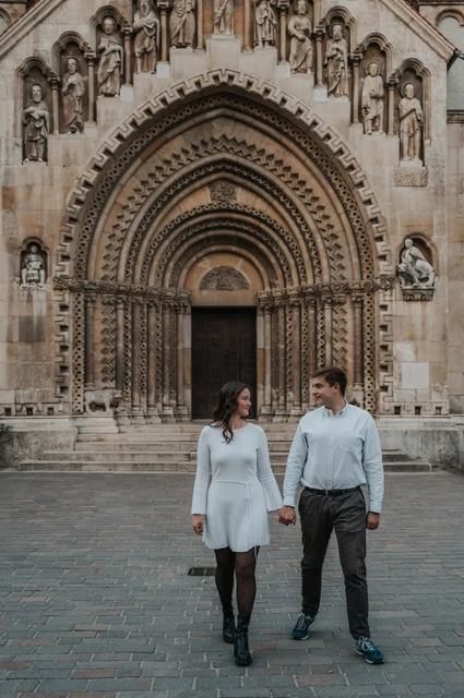 Walking together beside the castle chapel at a Budapest anniversary photoshoot The couple walking together in front of the chapel doorway at Vajdahunyad Castle during their anniversary photoshoot in Budapest.