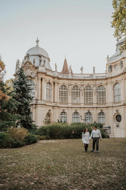 The grand palace façade behind the couple at a Budapest anniversary photoshoot A wide portrait of the couple standing on the grass beneath the ornate pale façade at Vajdahunyad Castle during their anniversary photoshoot in Budapest.
