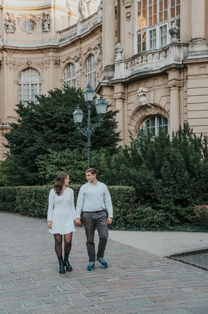 A walk beside the palace façade at a Budapest anniversary photoshoot The couple walking beside hedges and ornate lamps with Vajdahunyad Castle’s pale palace façade behind them during their anniversary photoshoot in Budapest.