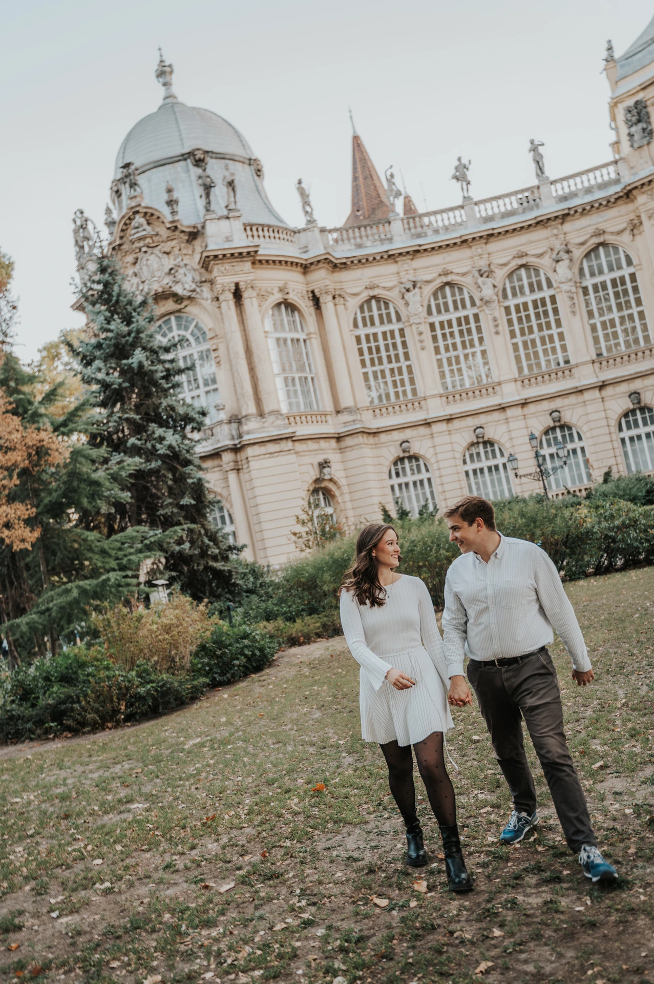 The couple holding hands on the lawn with Vajdahunyad Castle’s palace façade behind them during their anniversary photoshoot in Budapest.