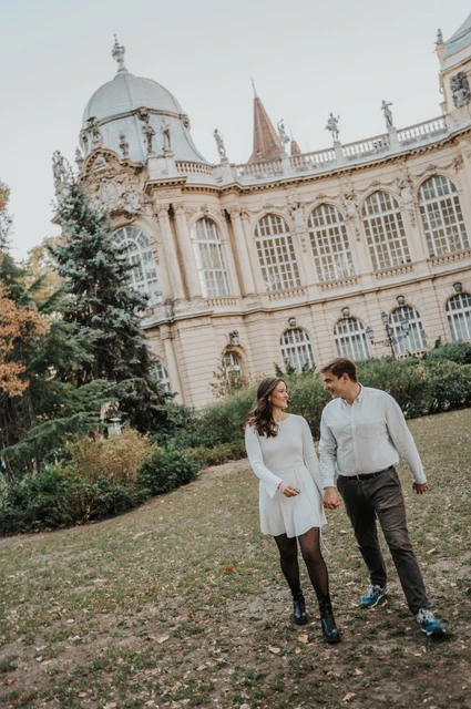 Holding hands in the castle gardens at a Budapest anniversary photoshoot The couple holding hands on the lawn with Vajdahunyad Castle’s palace façade behind them during their anniversary photoshoot in Budapest.