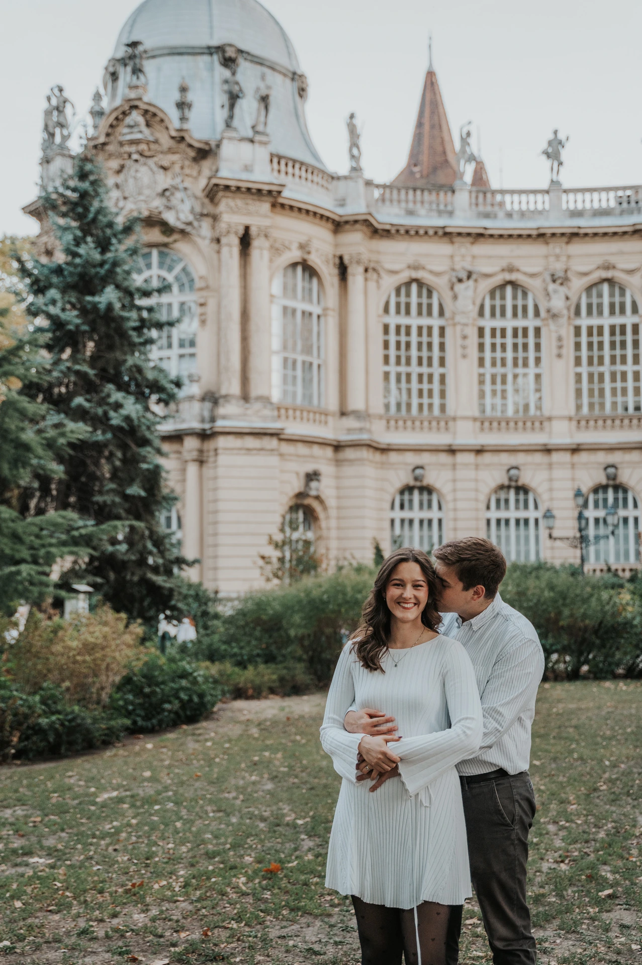 The man embracing his smiling partner from behind on the lawn in front of Vajdahunyad Castle during their anniversary photoshoot in Budapest.