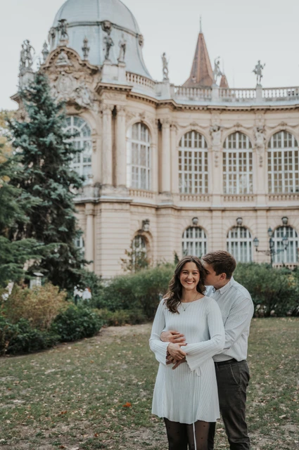 Smiles in front of the palace at a Budapest anniversary photoshoot The man embracing his smiling partner from behind on the lawn in front of Vajdahunyad Castle during their anniversary photoshoot in Budapest.
