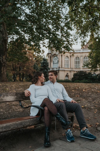 The couple sitting close together on a wooden bench beneath the trees near Vajdahunyad Castle during their anniversary photoshoot in Budapest.