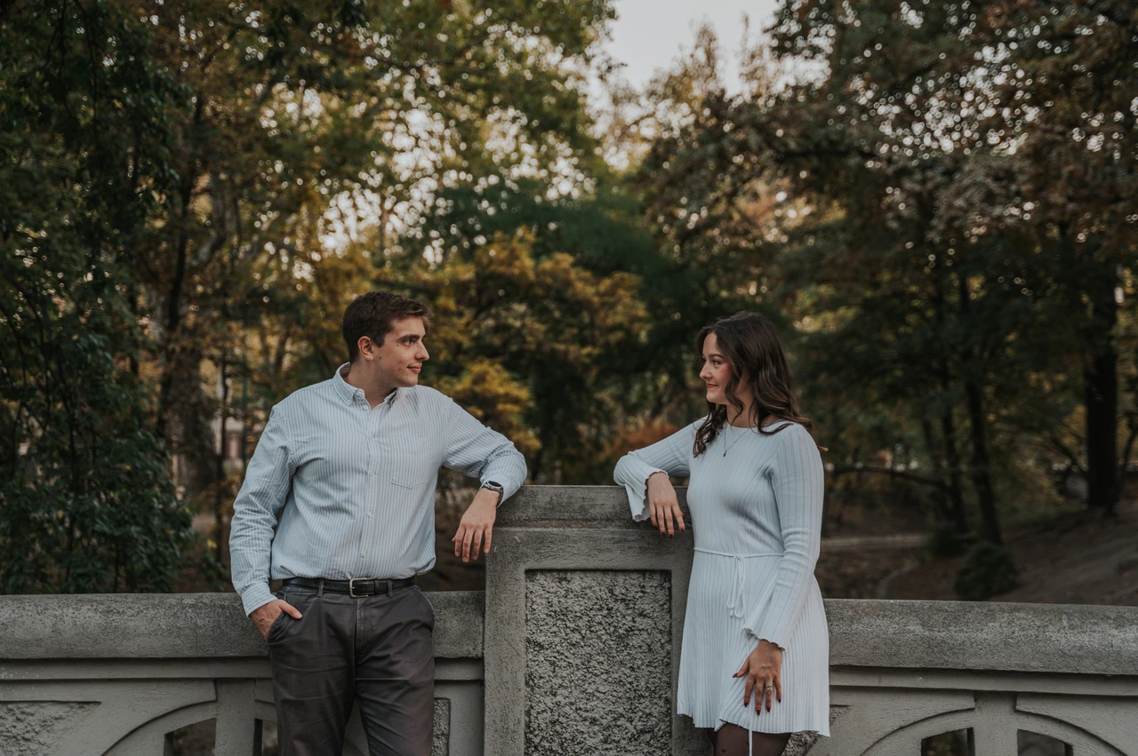 A relaxed portrait by the park bridge at a Budapest anniversary photoshoot A landscape portrait of the couple looking at one another beside a stone bridge railing near Vajdahunyad Castle during their anniversary photoshoot in Budapest.