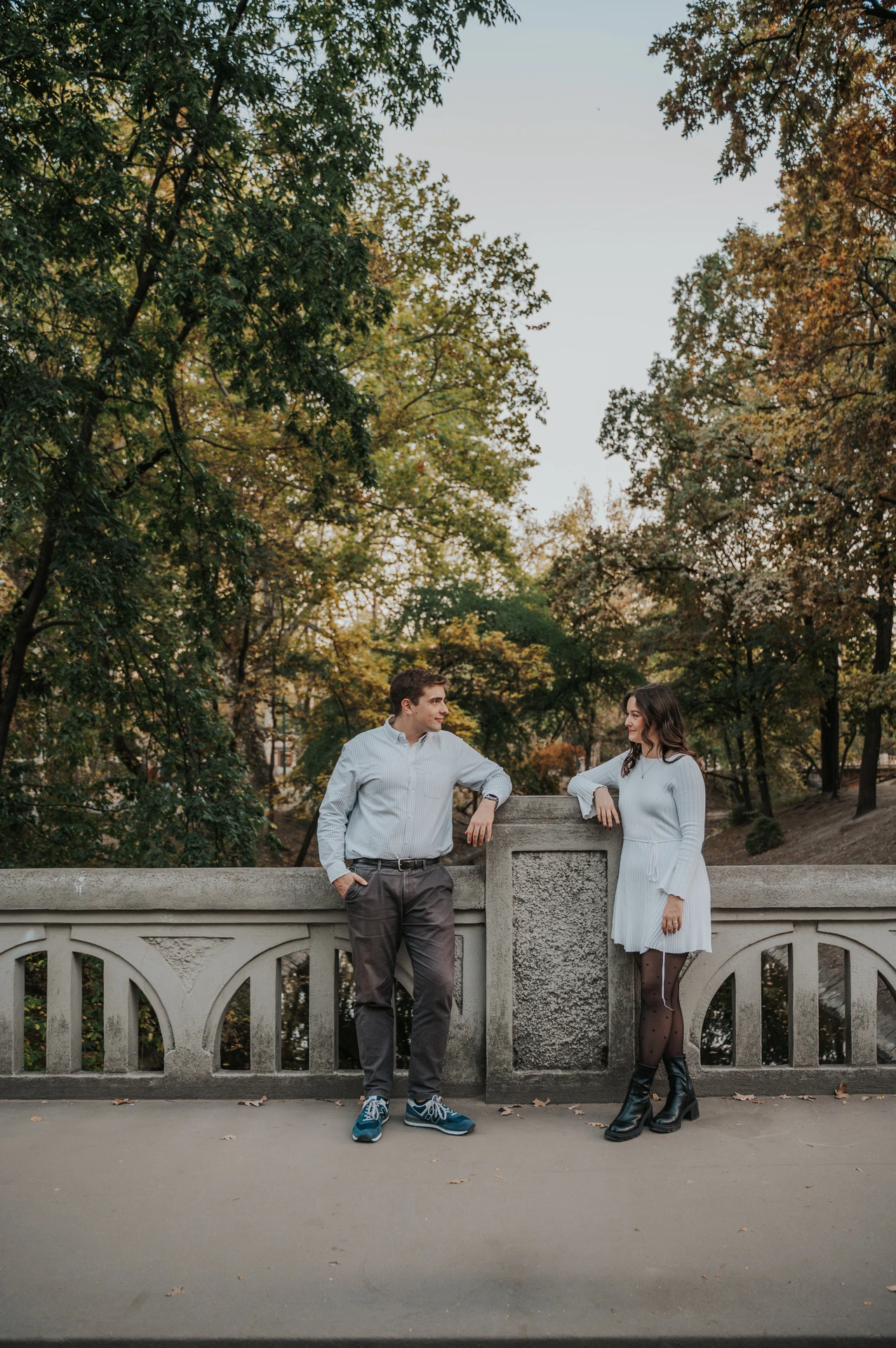 The couple leaning beside a stone bridge railing beneath leafy trees near Vajdahunyad Castle during their anniversary photoshoot in Budapest.