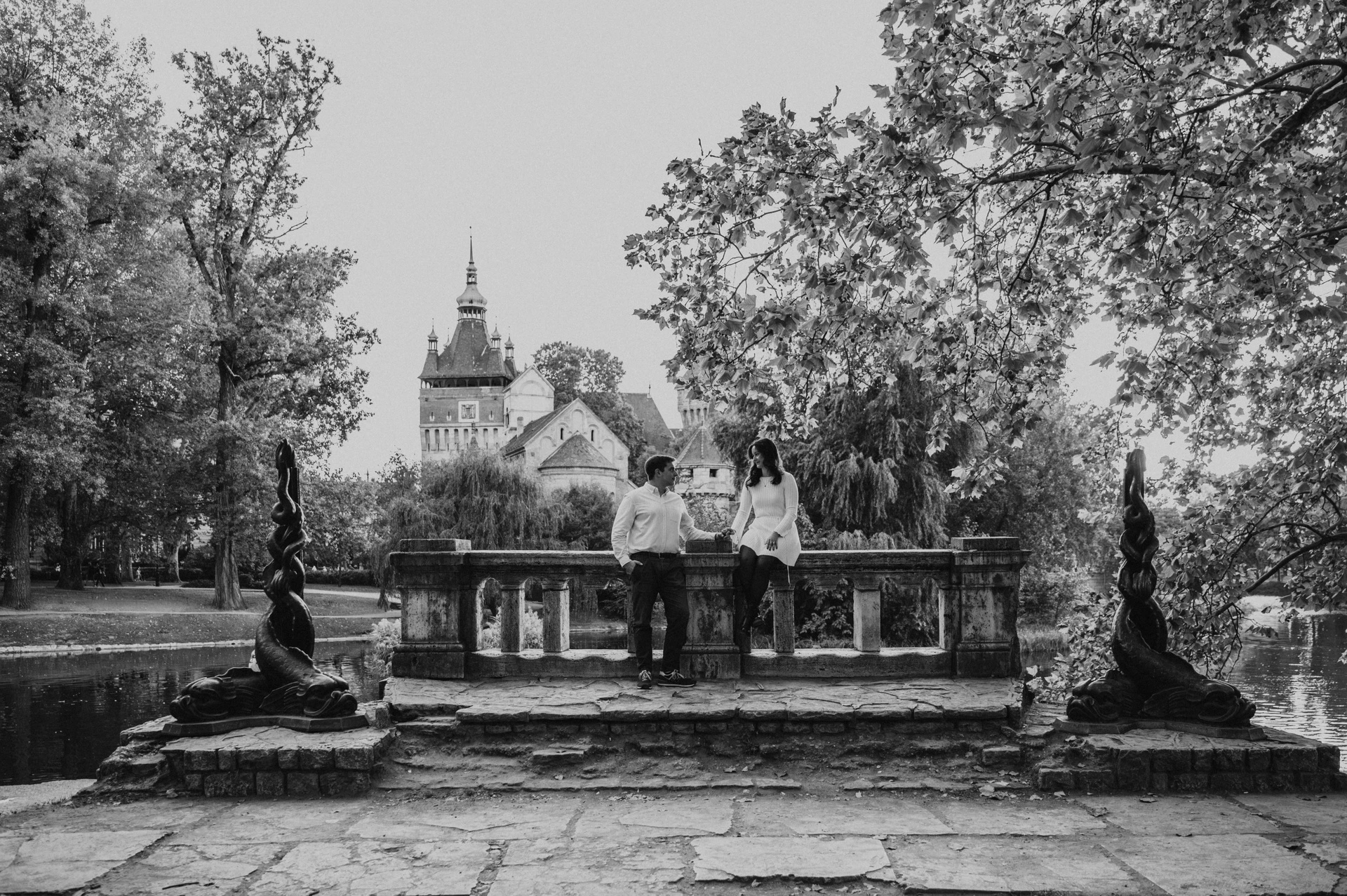 Black-and-white landscape portrait of the couple beside a stone balustrade framed by statues, trees and Vajdahunyad Castle during their anniversary photoshoot in Budapest.