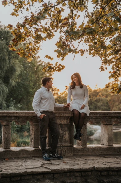 Sunlit leaves beside the water at a Budapest anniversary photoshoot The couple sitting beside each other on a stone balustrade beneath sunlit leaves near Vajdahunyad Castle during their anniversary photoshoot in Budapest.