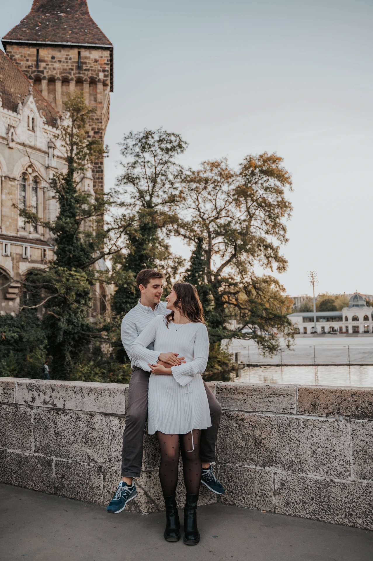 The couple embracing beside a low stone wall with Vajdahunyad Castle’s tower and palace façade behind them during their anniversary photoshoot in Budapest.