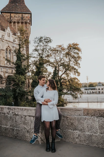 An embrace beside the castle tower at a Budapest anniversary photoshoot The couple embracing beside a low stone wall with Vajdahunyad Castle’s tower and palace façade behind them during their anniversary photoshoot in Budapest.