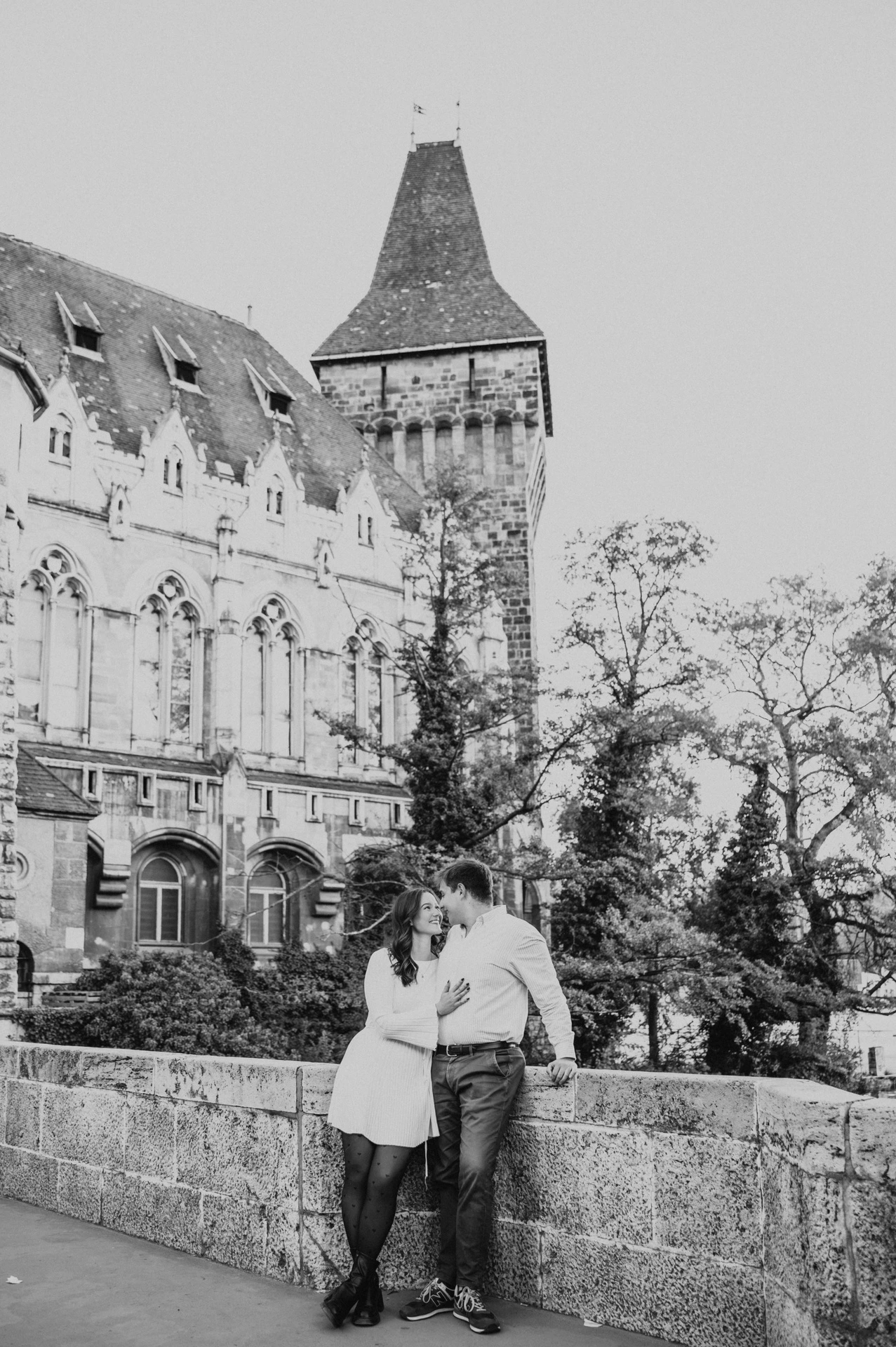 Black-and-white portrait of the couple leaning close beside a stone wall beneath Vajdahunyad Castle’s tower during their anniversary photoshoot in Budapest.