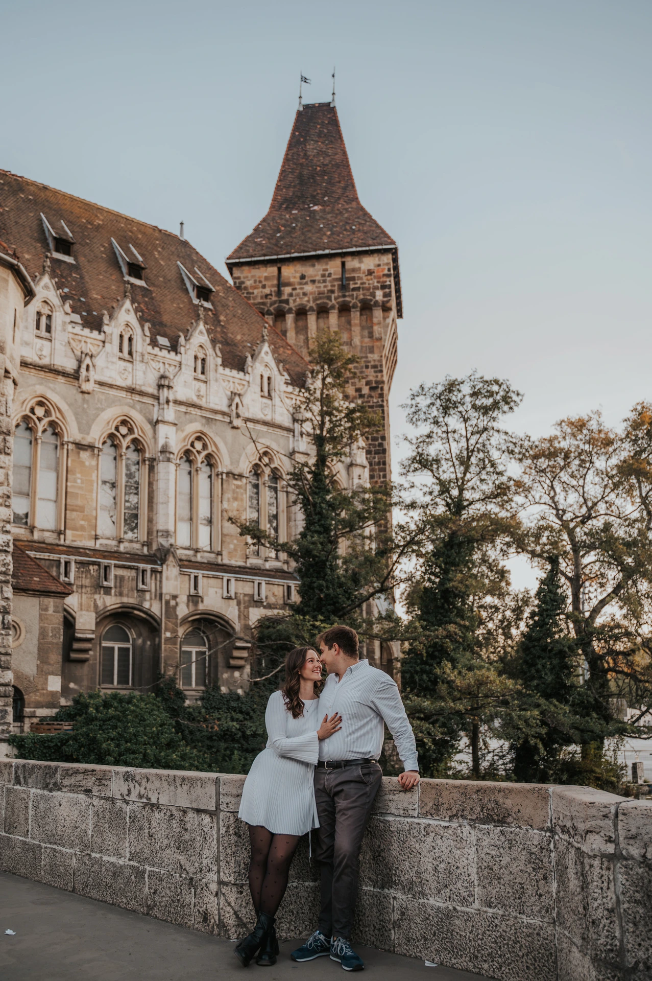 The couple leaning towards one another beside a stone wall beneath Vajdahunyad Castle’s tall tower during their anniversary photoshoot in Budapest.