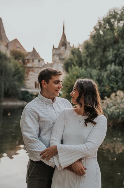 The couple smiling at one another beside the water with Vajdahunyad Castle’s turrets in the background during their anniversary photoshoot in Budapest.