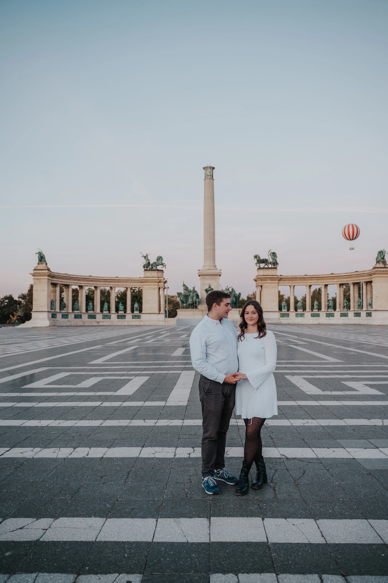 A full-length portrait of the couple at Heroes’ Square with the central column and curved colonnade behind them during their anniversary photoshoot in Budapest.