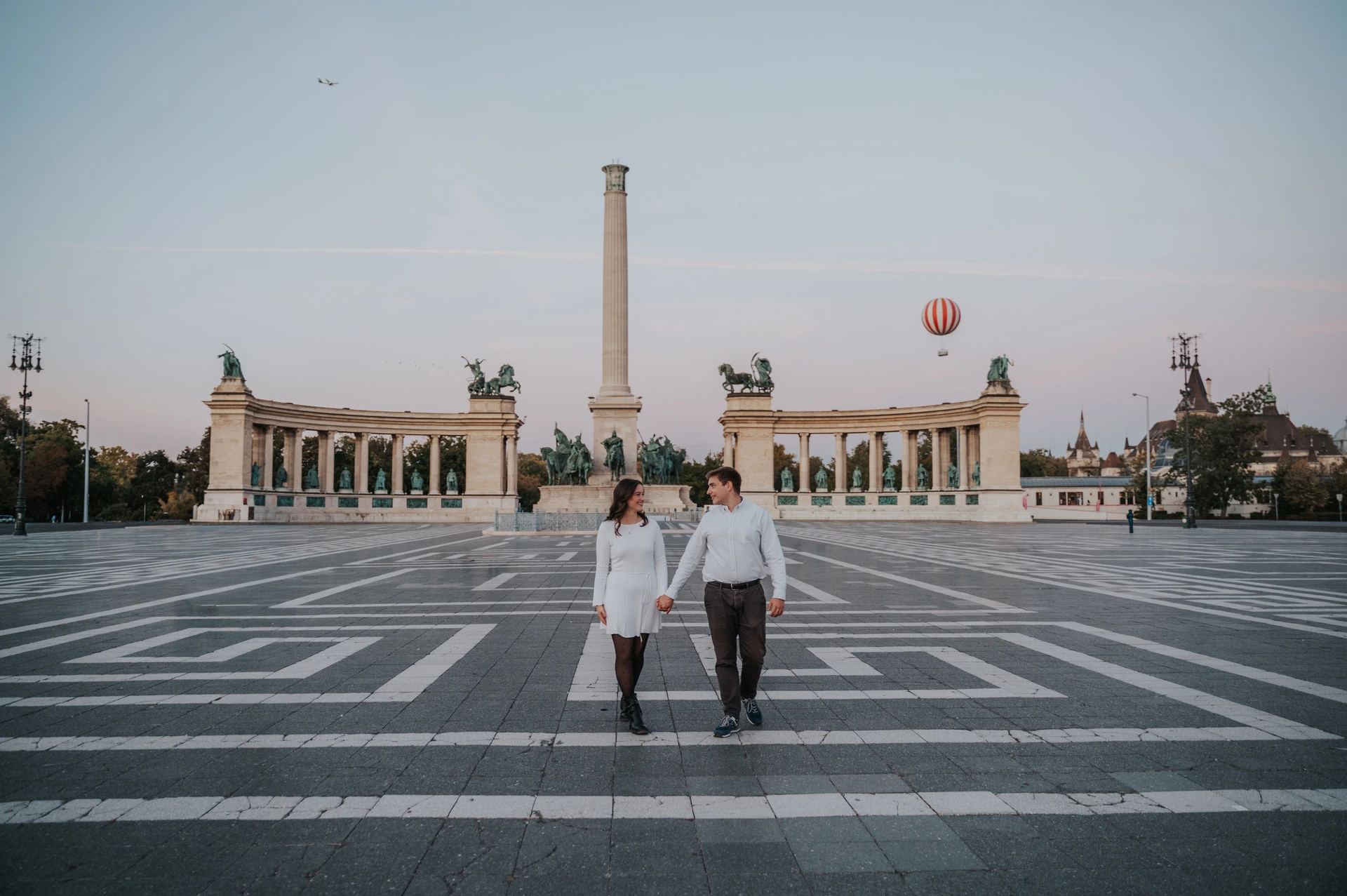 A wide landscape portrait of the couple walking across the patterned paving with Heroes’ Square’s column and colonnade behind them during their anniversary photoshoot in Budapest.