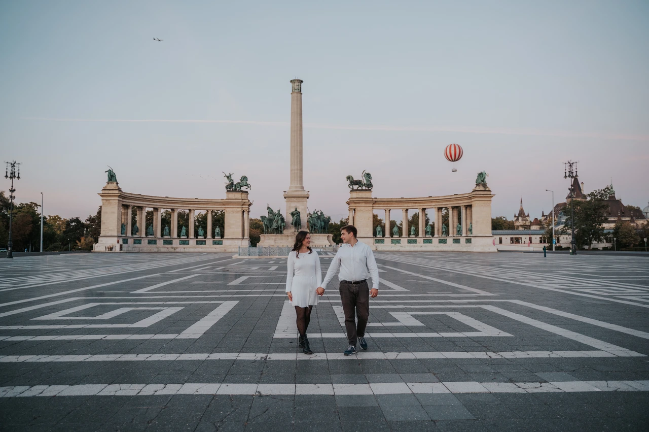 A walk across the grand square at a Budapest anniversary photoshoot A wide landscape portrait of the couple walking across the patterned paving with Heroes’ Square’s column and colonnade behind them during their anniversary photoshoot in Budapest.