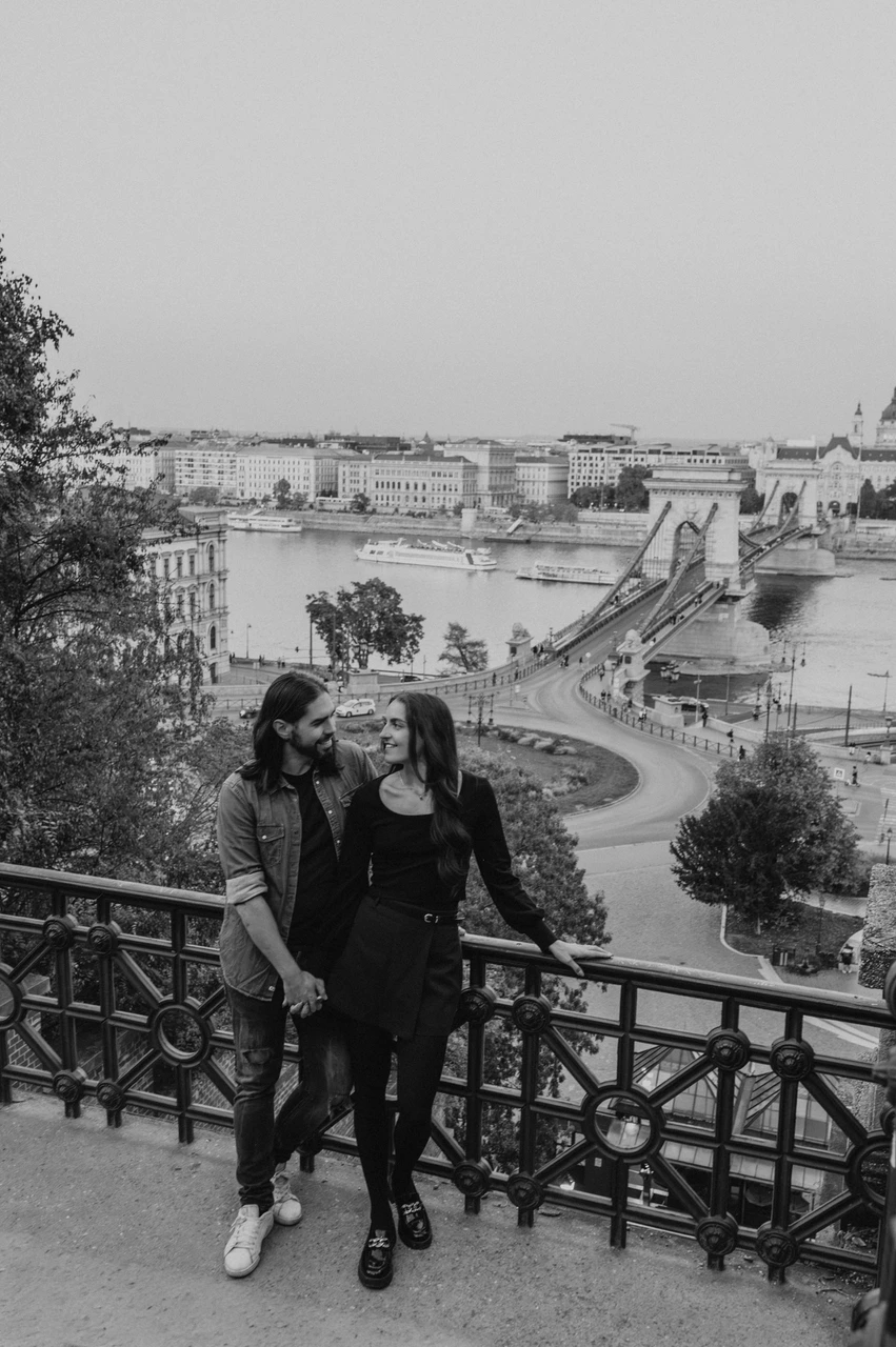 The Chain Bridge below in black and white at a Budapest couple photoshoot Black-and-white portrait of the couple beside a decorative railing with the Chain Bridge and Danube behind them during their couple photoshoot in Budapest.