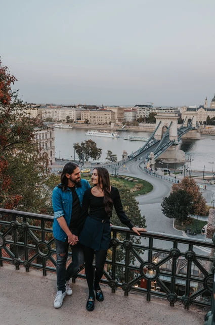 The couple standing beside a decorative railing with the Chain Bridge and Danube below Buda Castle during their couple photoshoot in Budapest.