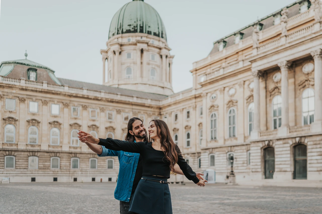 The palace courtyard and green dome at a Budapest couple photoshoot A landscape portrait of the couple with outstretched arms in the courtyard beneath Buda Castle’s green dome during their couple photoshoot in Budapest.