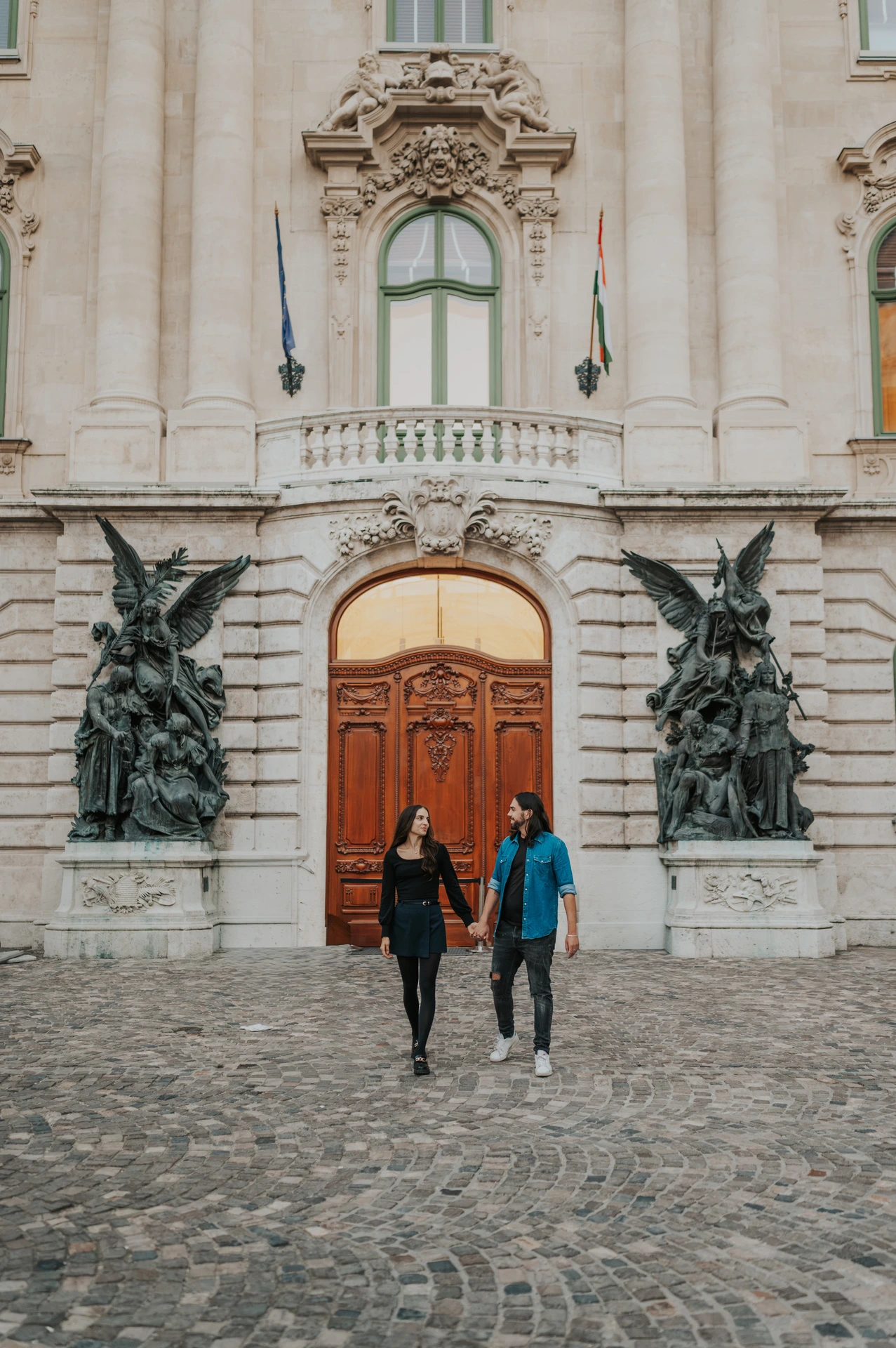 The couple walking across the cobbled courtyard with an ornate wooden door and bronze sculptures at Buda Castle behind them during their couple photoshoot in Budapest.