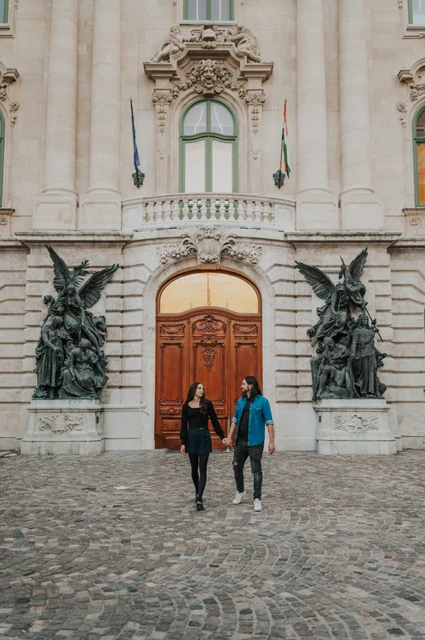 A walk across the castle courtyard at a Budapest couple photoshoot The couple walking across the cobbled courtyard with an ornate wooden door and bronze sculptures at Buda Castle behind them during their couple photoshoot in Budapest.