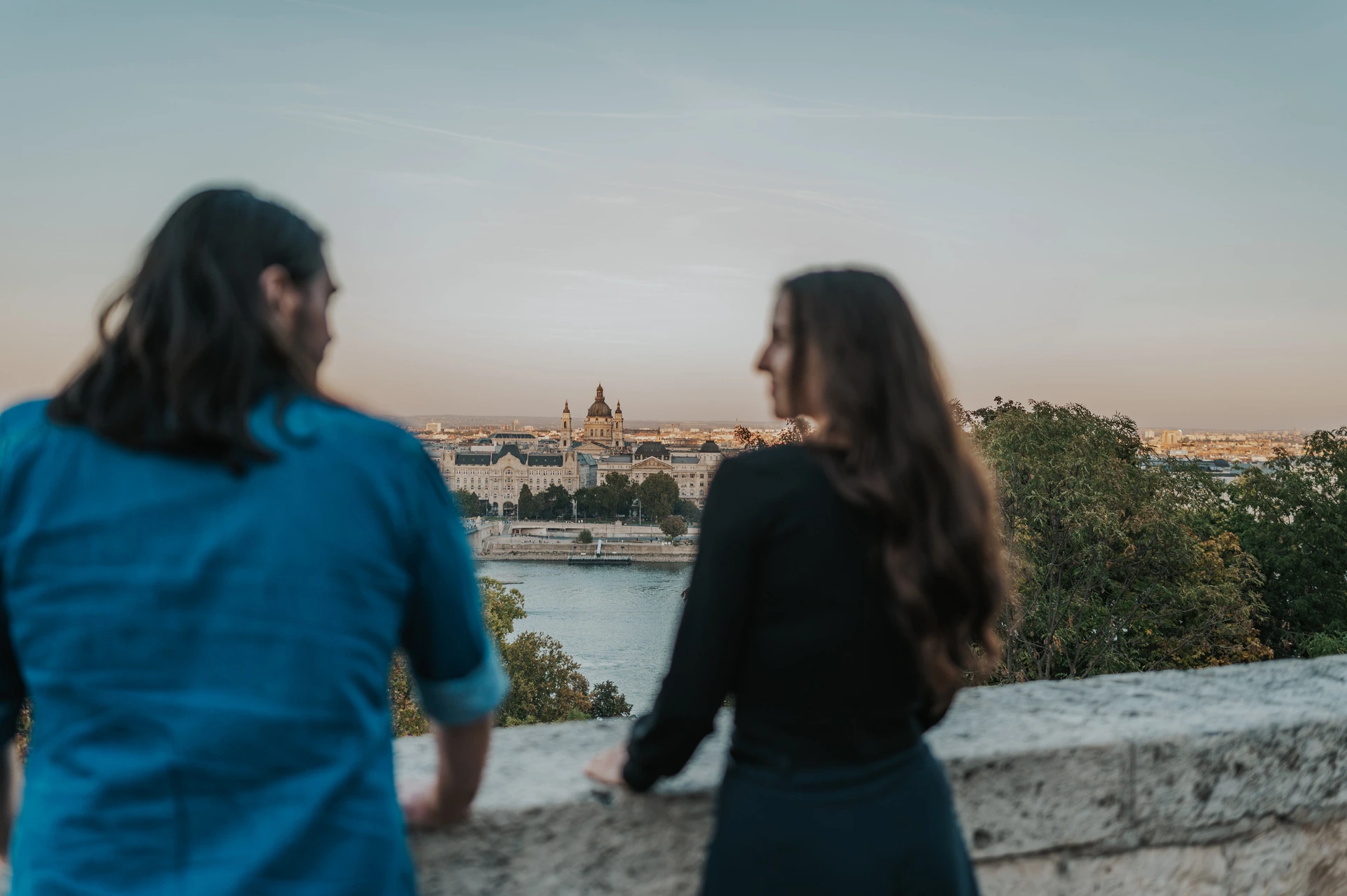 The couple seen from behind looking across the Danube from a stone terrace at Buda Castle during their couple photoshoot in Budapest.
