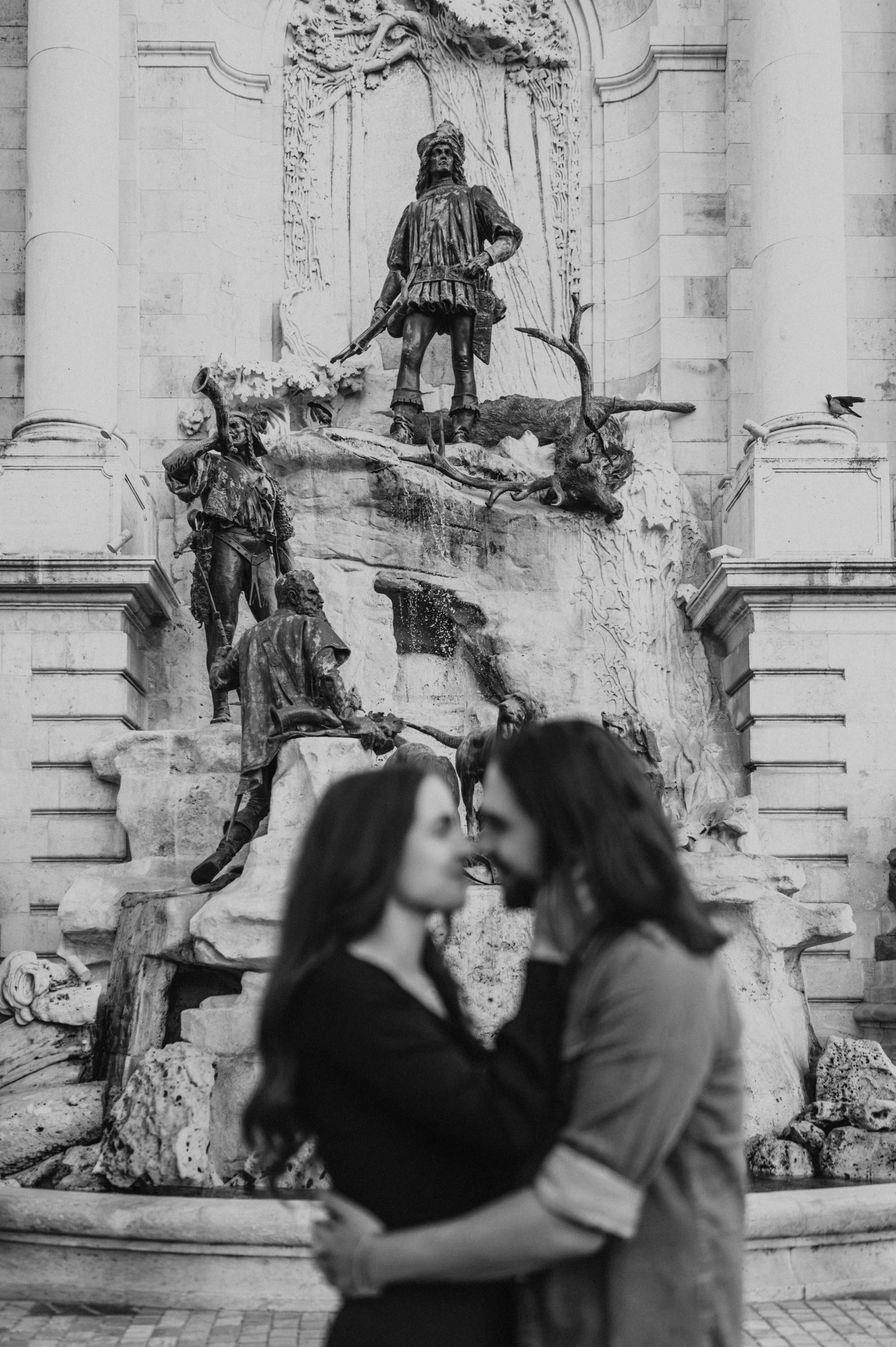 Black-and-white portrait of the couple embracing in front of the bronze figures at Buda Castle’s fountain during their couple photoshoot in Budapest.