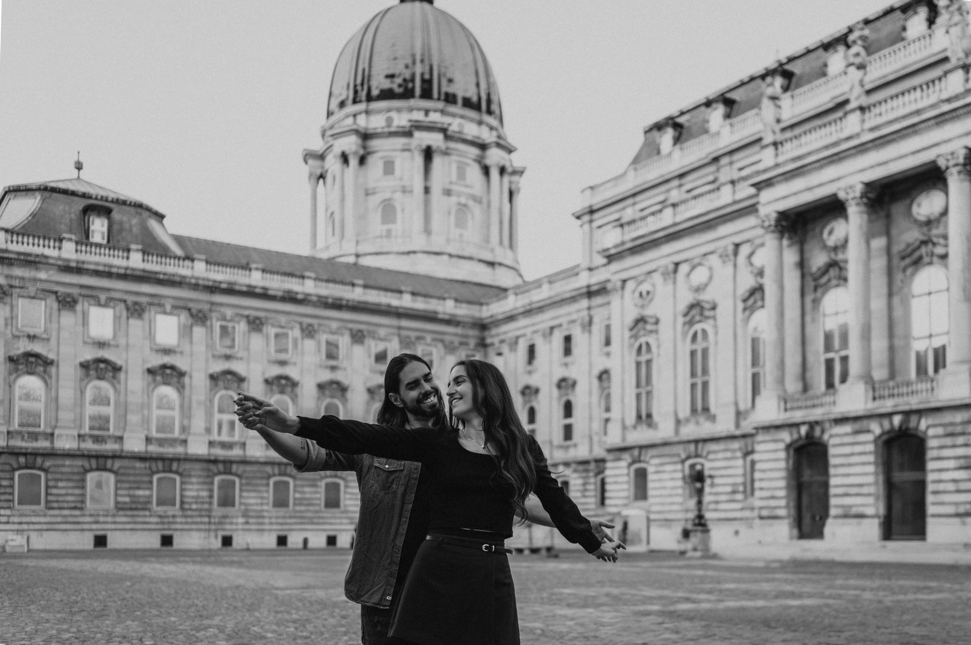 Black-and-white landscape portrait of the couple with outstretched arms beneath Buda Castle’s green dome during their couple photoshoot in Budapest.