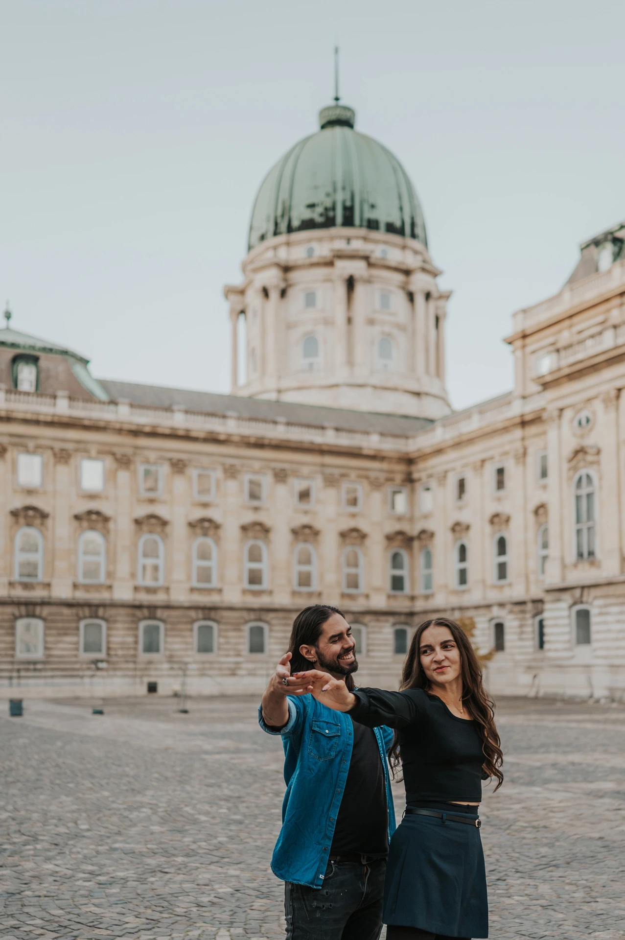 The couple sharing a playful portrait with outstretched arms beneath Buda Castle’s green dome during their couple photoshoot in Budapest.