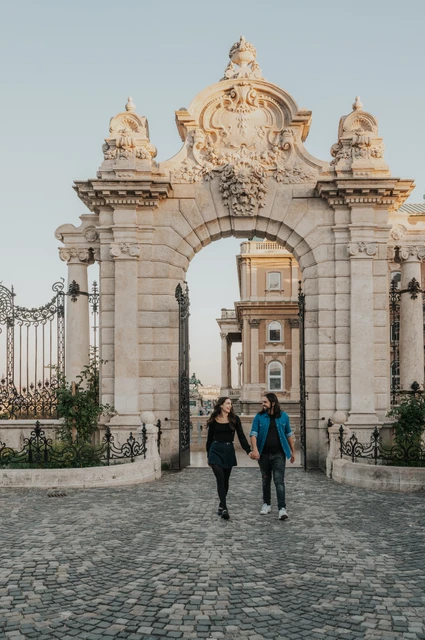 The couple walking hand in hand through an ornate stone gateway at Buda Castle during their couple photoshoot in Budapest.