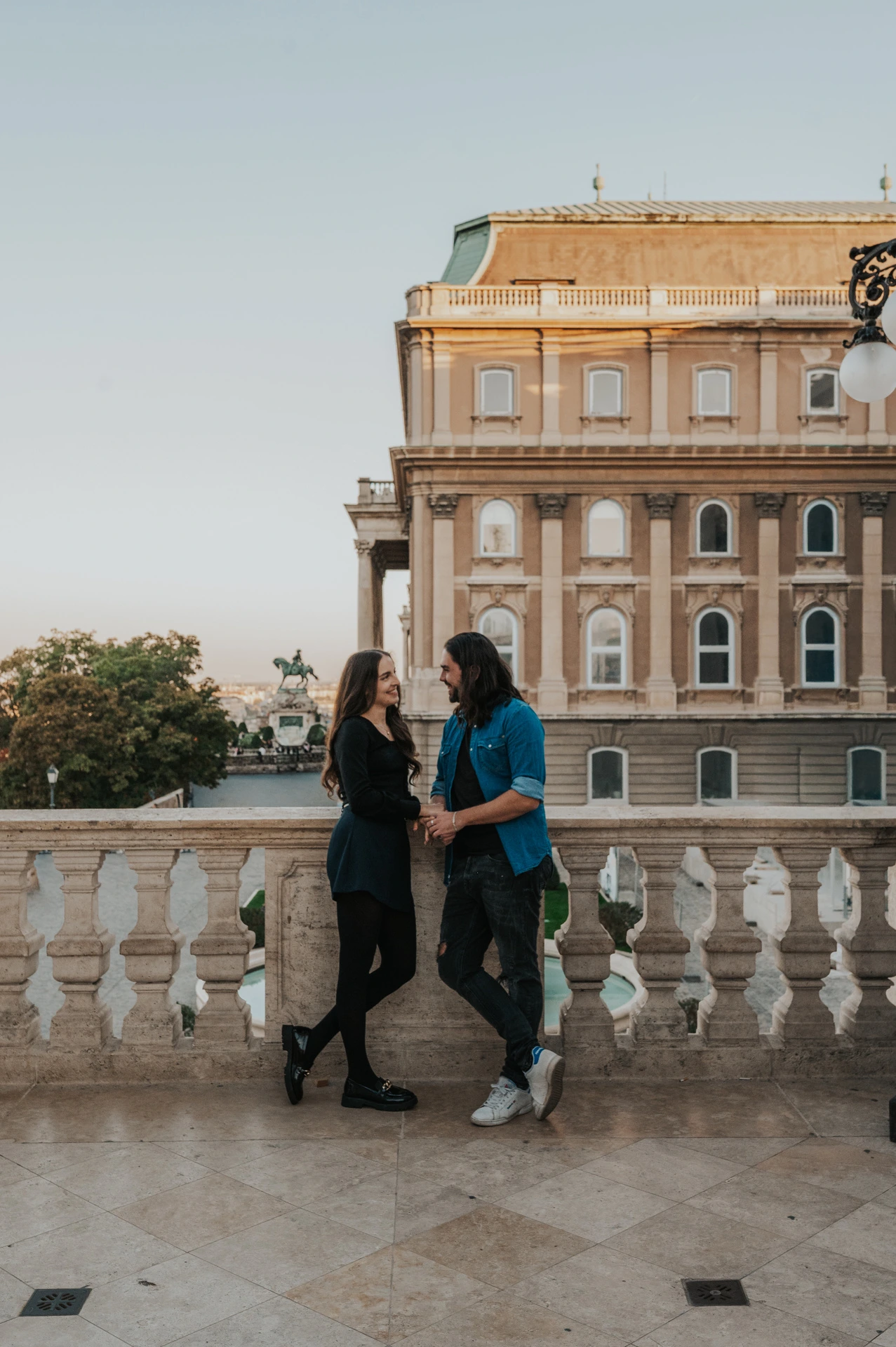 The couple walking together beside a stone balustrade with Buda Castle’s palace façade behind them during their couple photoshoot in Budapest.