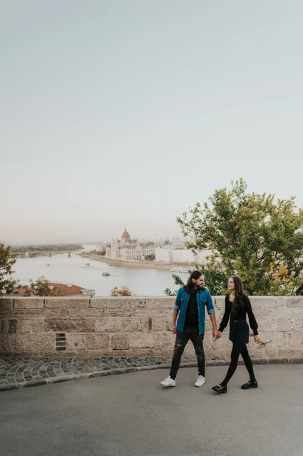 Walking beside the city viewpoint at a Budapest couple photoshoot The couple walking hand in hand beside the stone wall with the Danube and Budapest skyline beyond Buda Castle during their couple photoshoot in Budapest.