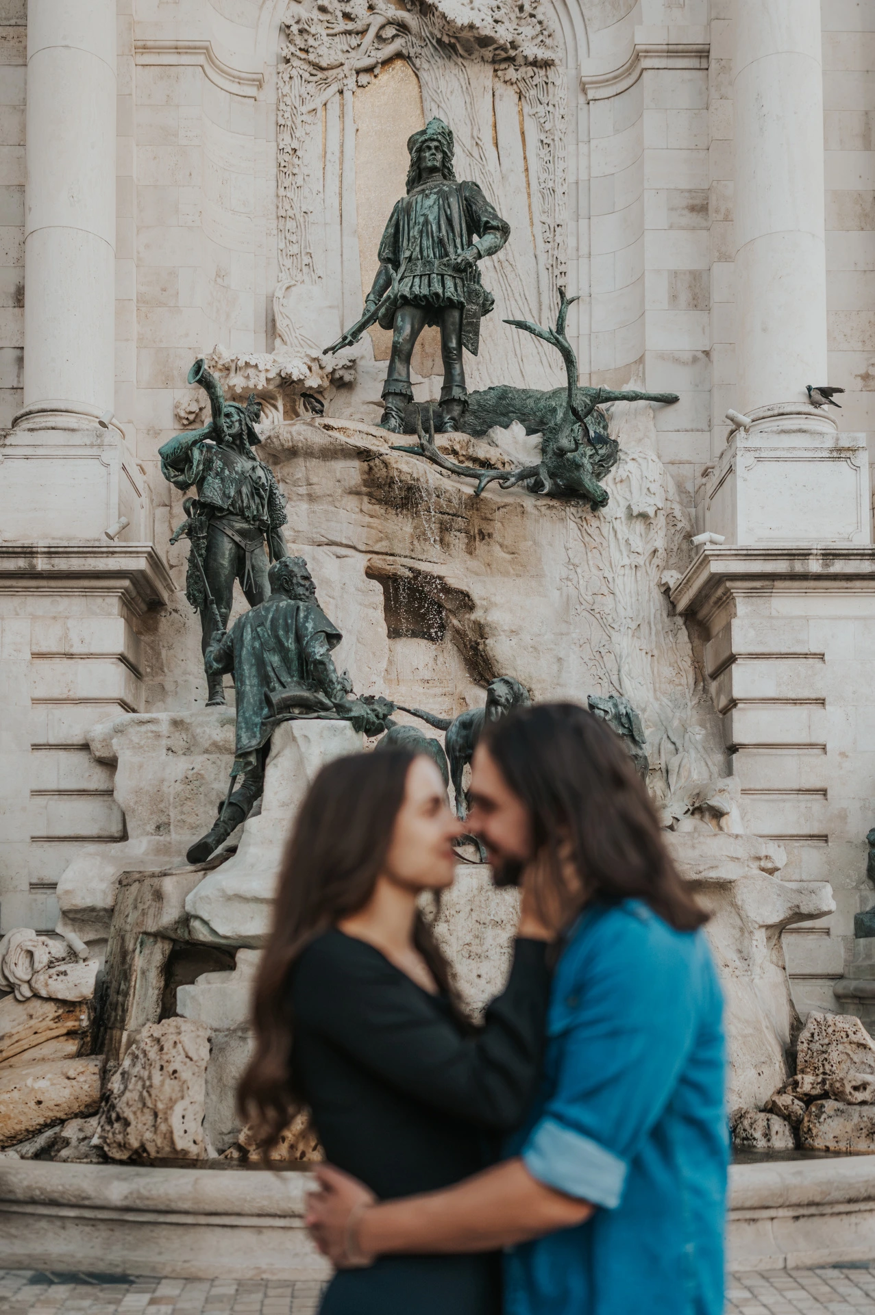 The couple embracing in front of Buda Castle’s sculpted stone fountain and bronze figures during their couple photoshoot in Budapest.