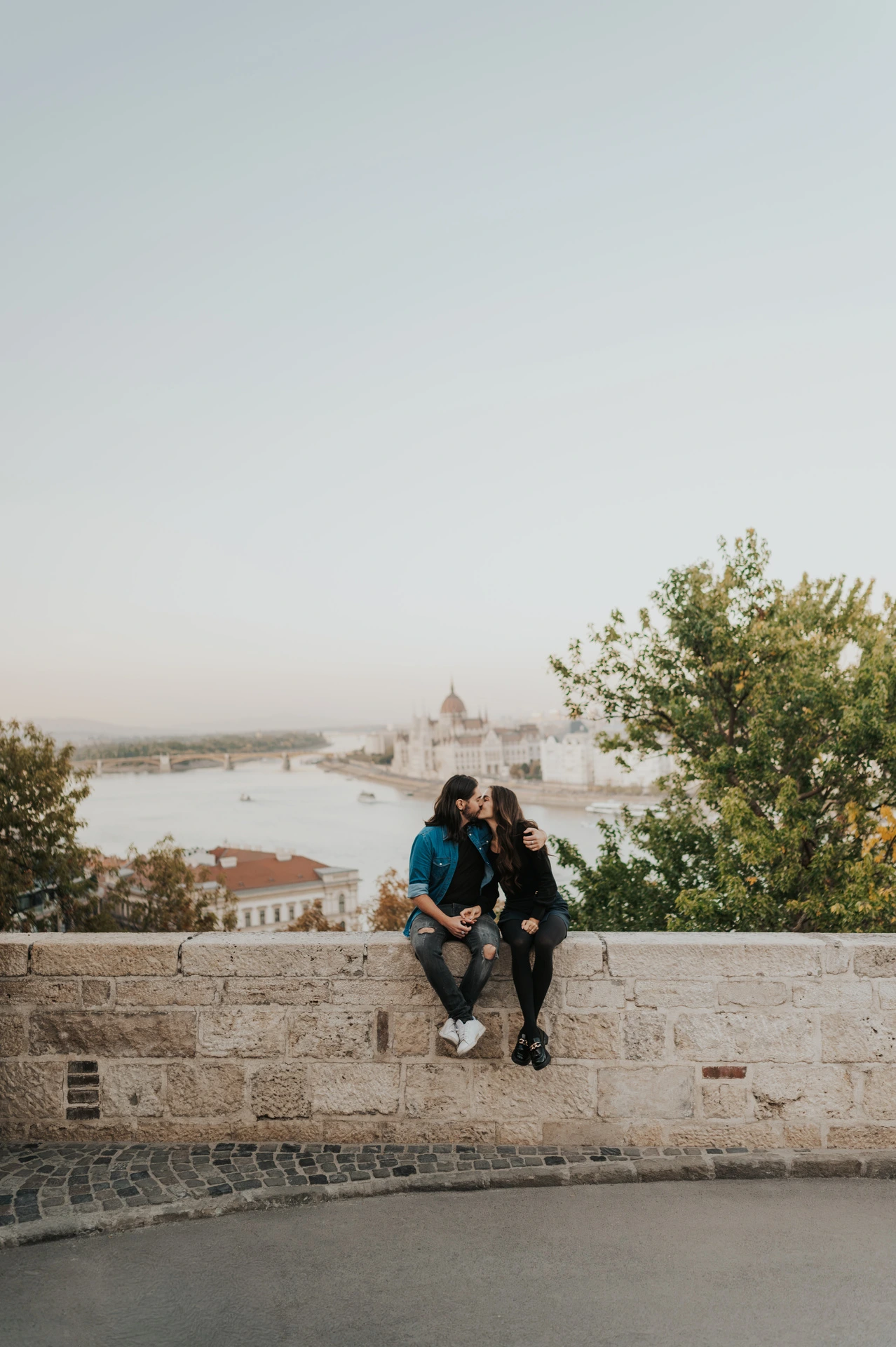 The couple sitting together on a low stone wall with the Danube and Budapest skyline behind them at Buda Castle during their couple photoshoot in Budapest.