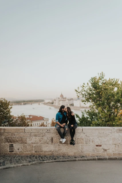 Sitting together above the Danube at a Budapest couple photoshoot The couple sitting together on a low stone wall with the Danube and Budapest skyline behind them at Buda Castle during their couple photoshoot in Budapest.