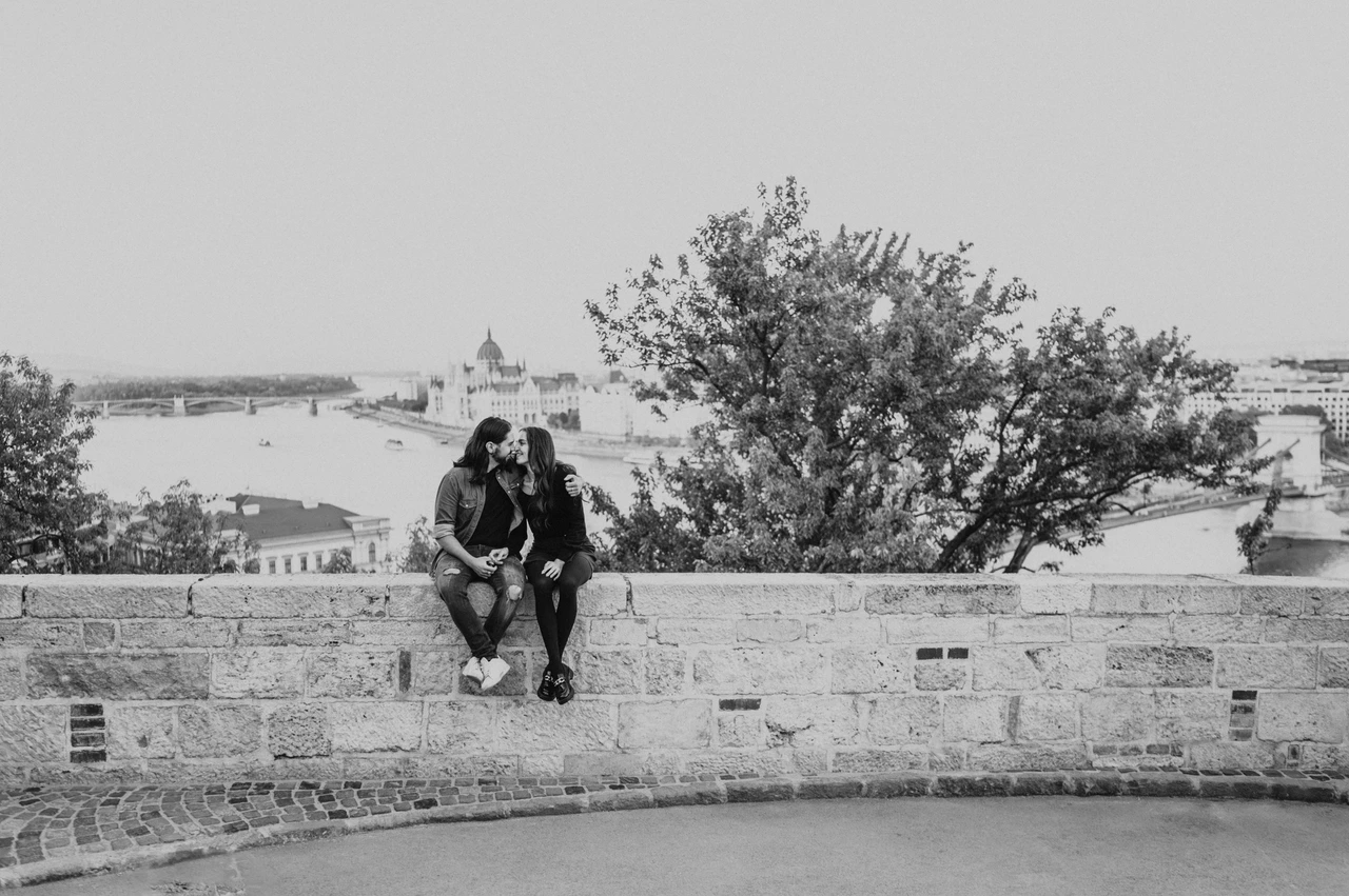 A wide riverside portrait in black and white at a Budapest couple photoshoot Black-and-white landscape portrait of the couple sitting on a stone wall with the Danube and Budapest skyline behind them during their couple photoshoot in Budapest.