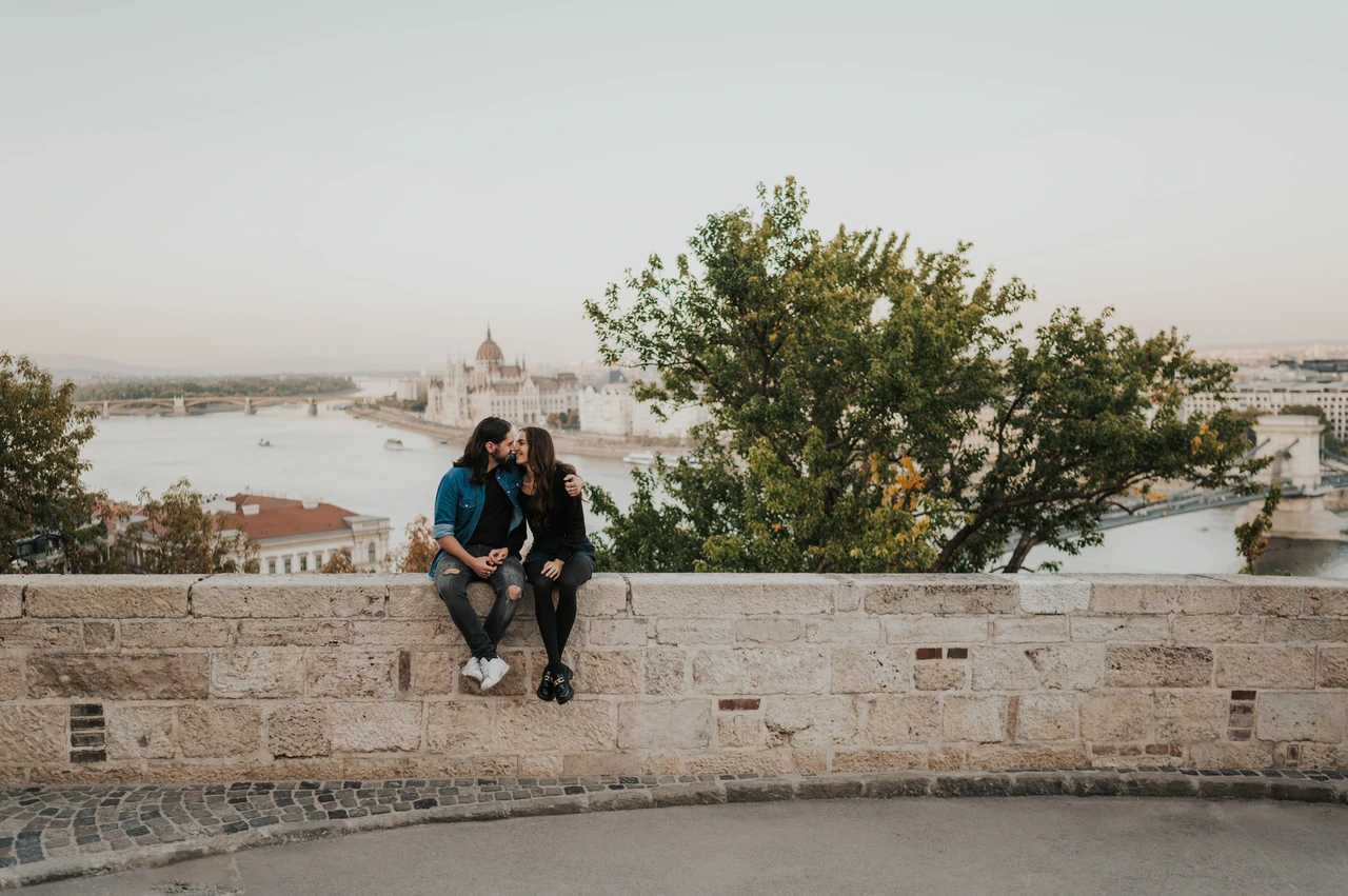 A wide view of the city and river at a Budapest couple photoshoot A wide landscape portrait of the couple sitting on a stone wall overlooking the Danube from Buda Castle during their couple photoshoot in Budapest.