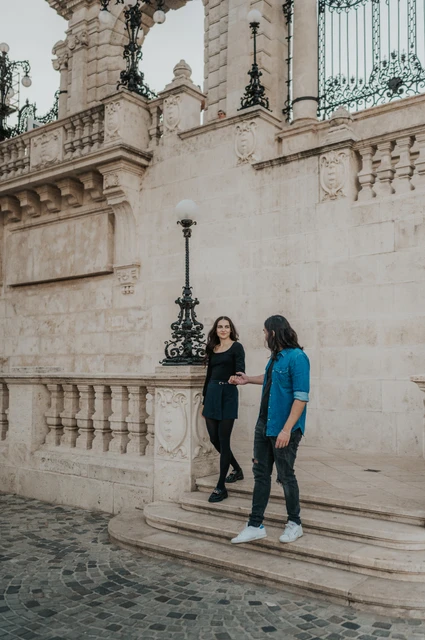 A walk down the castle steps at a Budapest couple photoshoot The couple walking down stone steps beside an ornate lamp and the pale walls at Buda Castle during their couple photoshoot in Budapest.