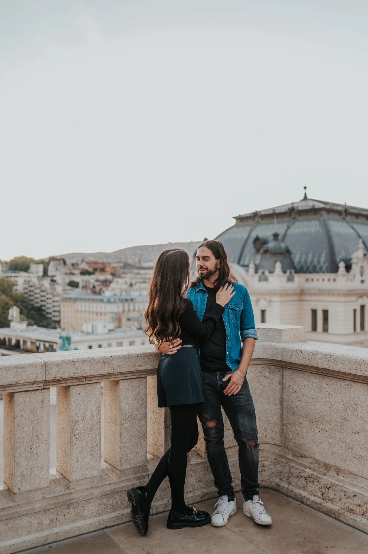 The couple standing close in the corner of a stone terrace at Buda Castle with city rooftops behind them during their couple photoshoot in Budapest.