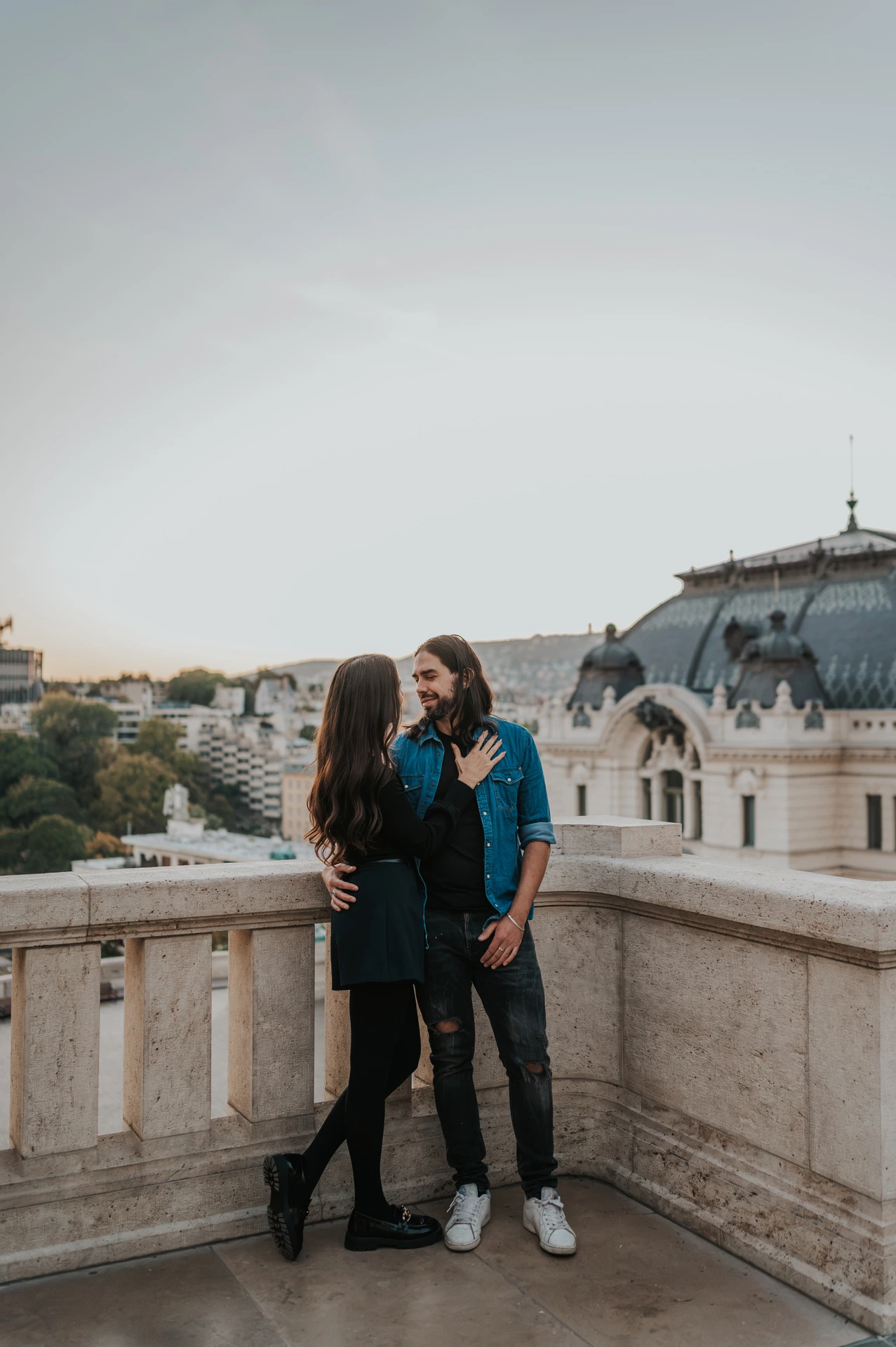 A full-length portrait of the couple embracing beside the corner of a stone terrace at Buda Castle during their couple photoshoot in Budapest.
