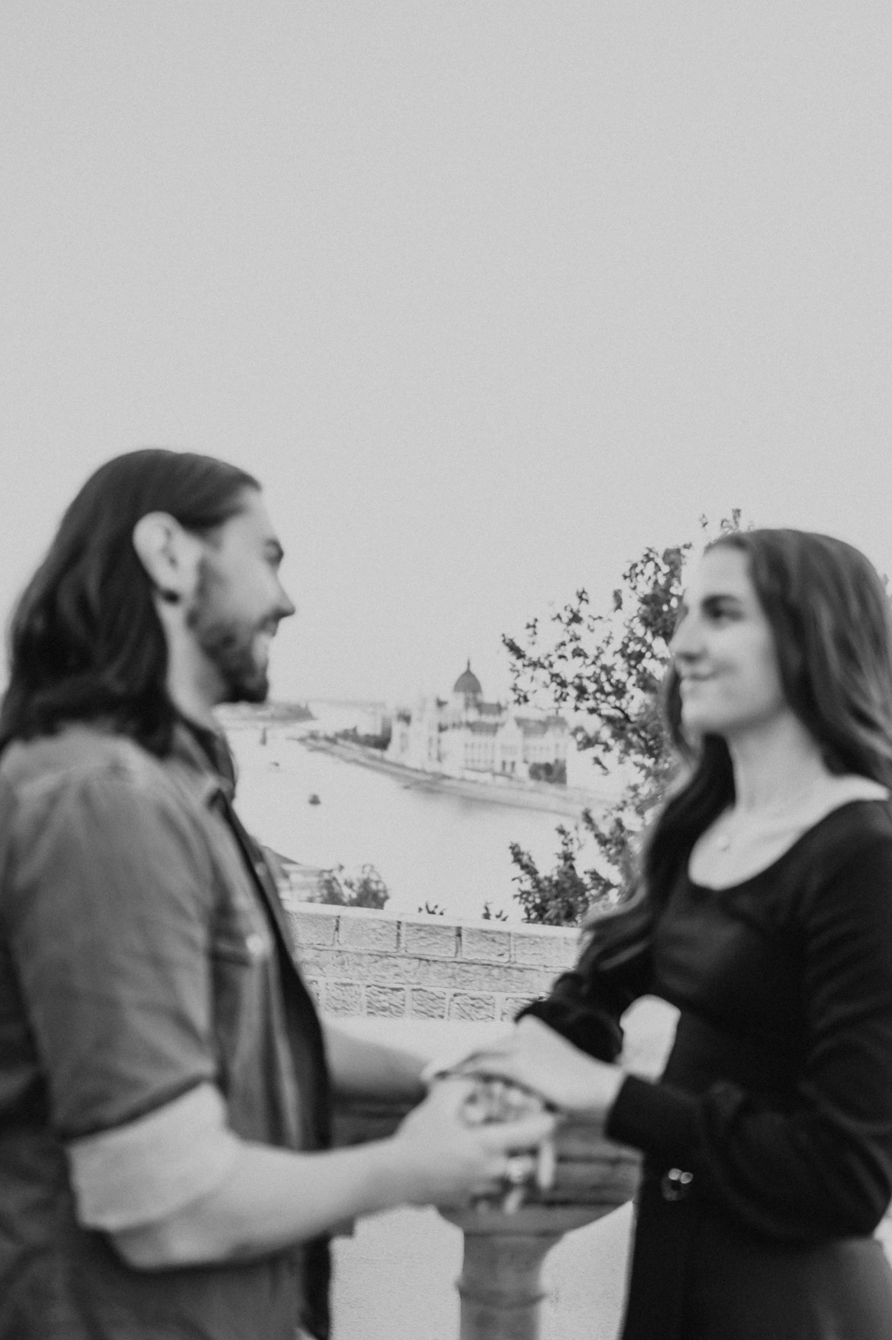 Black-and-white portrait of the couple holding hands on a terrace overlooking Budapest from Buda Castle during their couple photoshoot in Budapest.