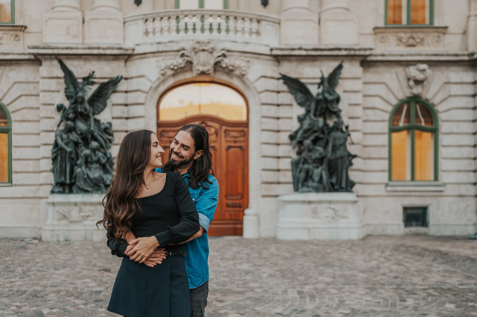 Smiles in front of the ornate palace door at a Budapest couple photoshoot A landscape portrait of the man embracing his smiling partner from behind in front of an ornate wooden door at Buda Castle during their couple photoshoot in Budapest.