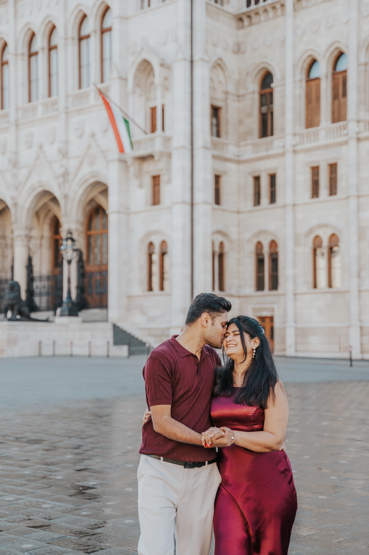 The couple touching foreheads in burgundy outfits in front of Parliament’s arched windows during their Budapest couple photoshoot.