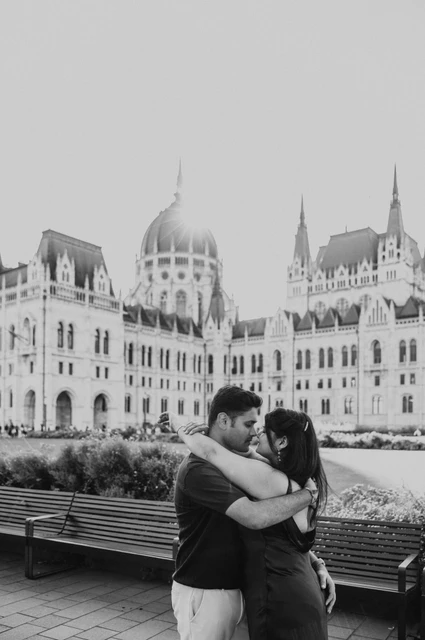 Budapest couple photoshoot on a bench in black and white Black-and-white portrait of the couple embracing on a bench with Parliament behind them during their couple photoshoot in Budapest.