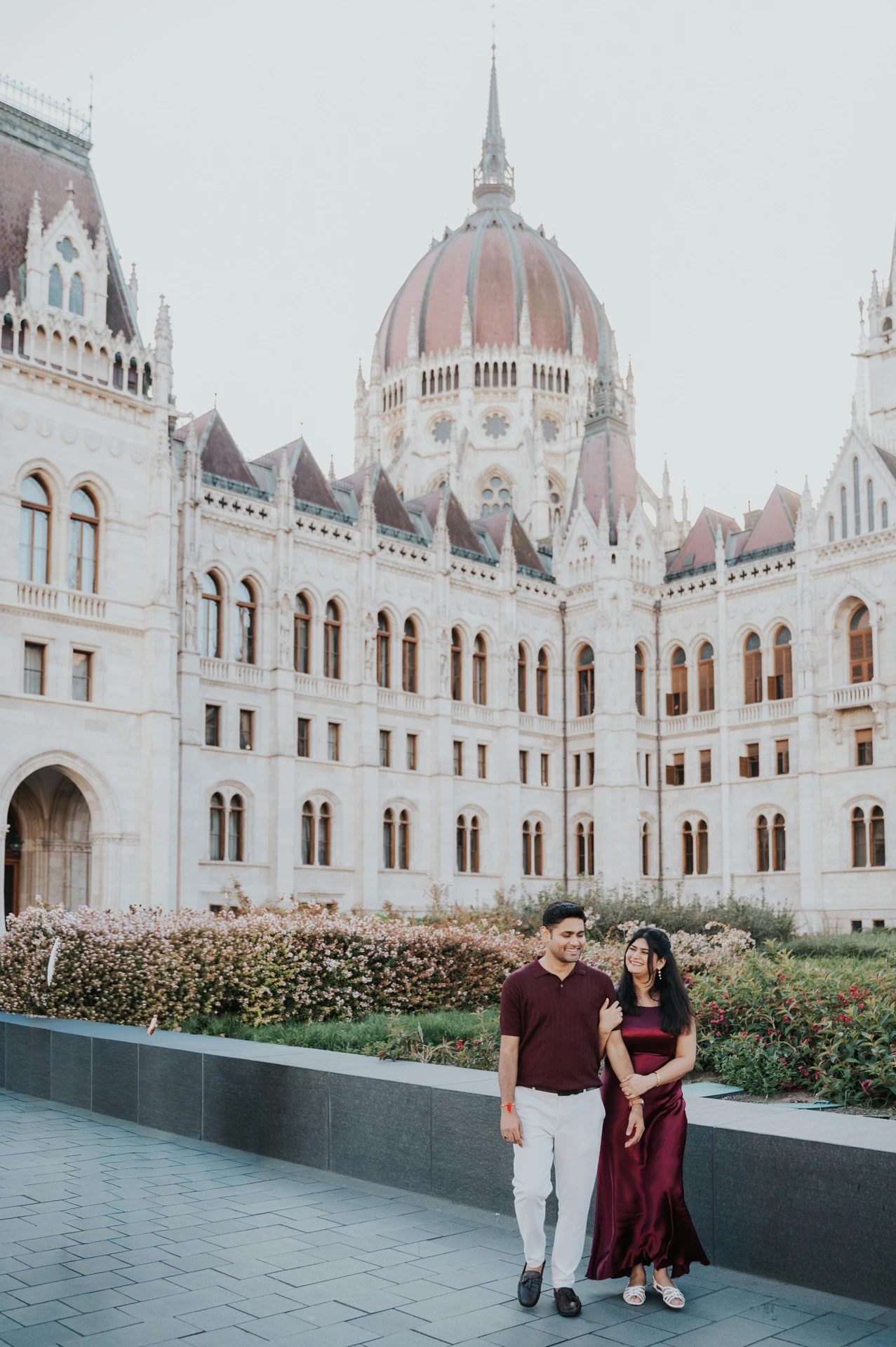 The couple standing hand in hand beside the flowerbeds with Parliament’s dome above them during their couple photoshoot in Budapest.