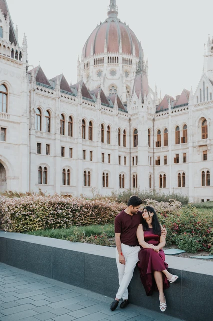 A seated portrait by Parliament at a Budapest couple photoshoot The couple sitting together beside flowerbeds with Parliament’s dome behind them during their Budapest couple photoshoot.