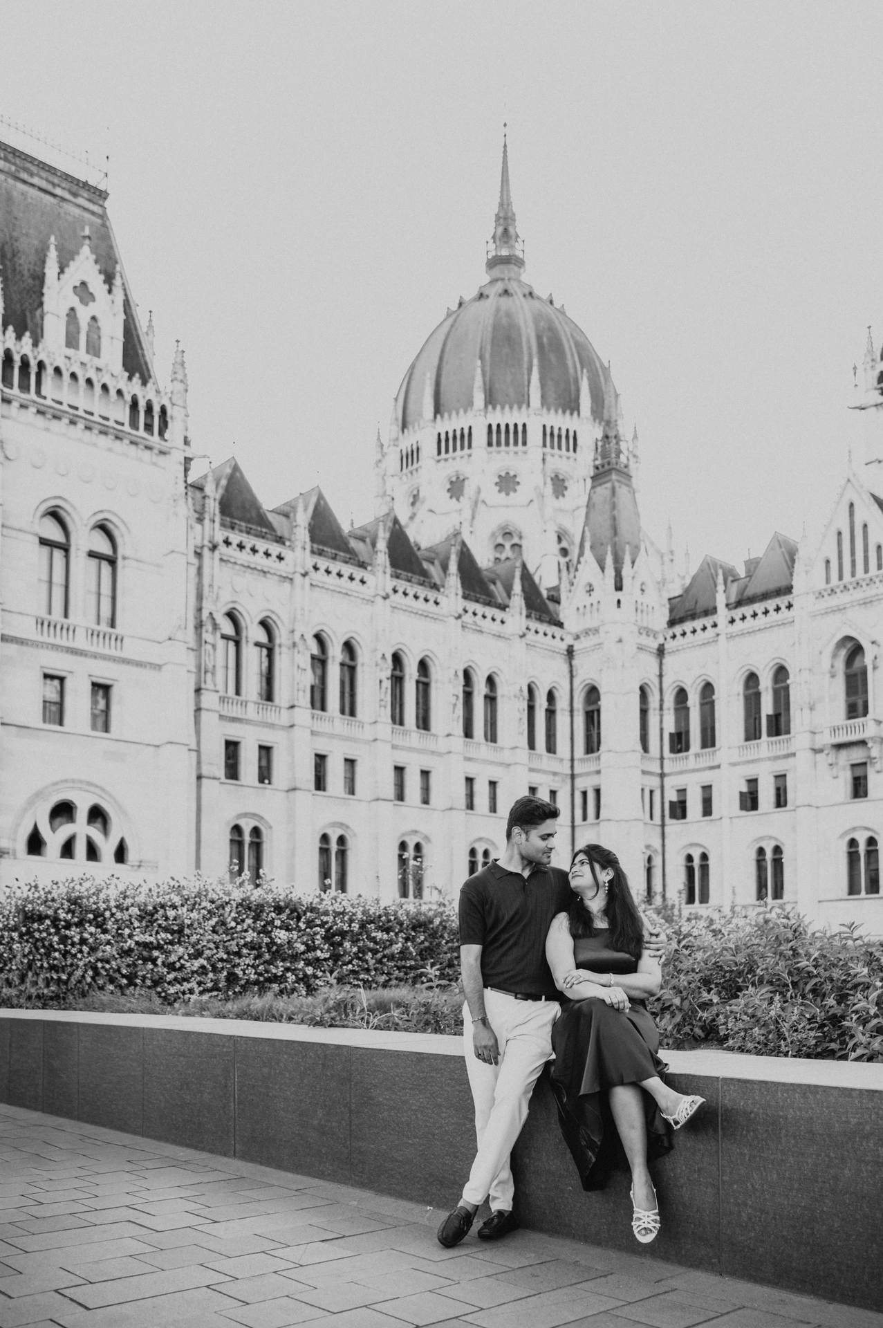 Black-and-white portrait of the couple sitting on a low wall beneath Parliament’s dome during their couple photoshoot in Budapest.