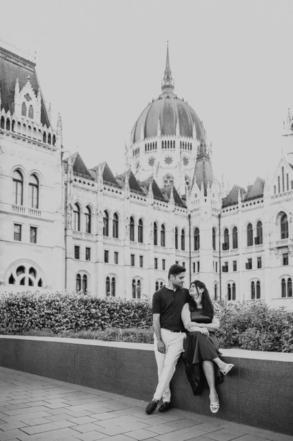 Budapest couple photoshoot beneath Parliament’s dome in black and white Black-and-white portrait of the couple sitting on a low wall beneath Parliament’s dome during their couple photoshoot in Budapest.