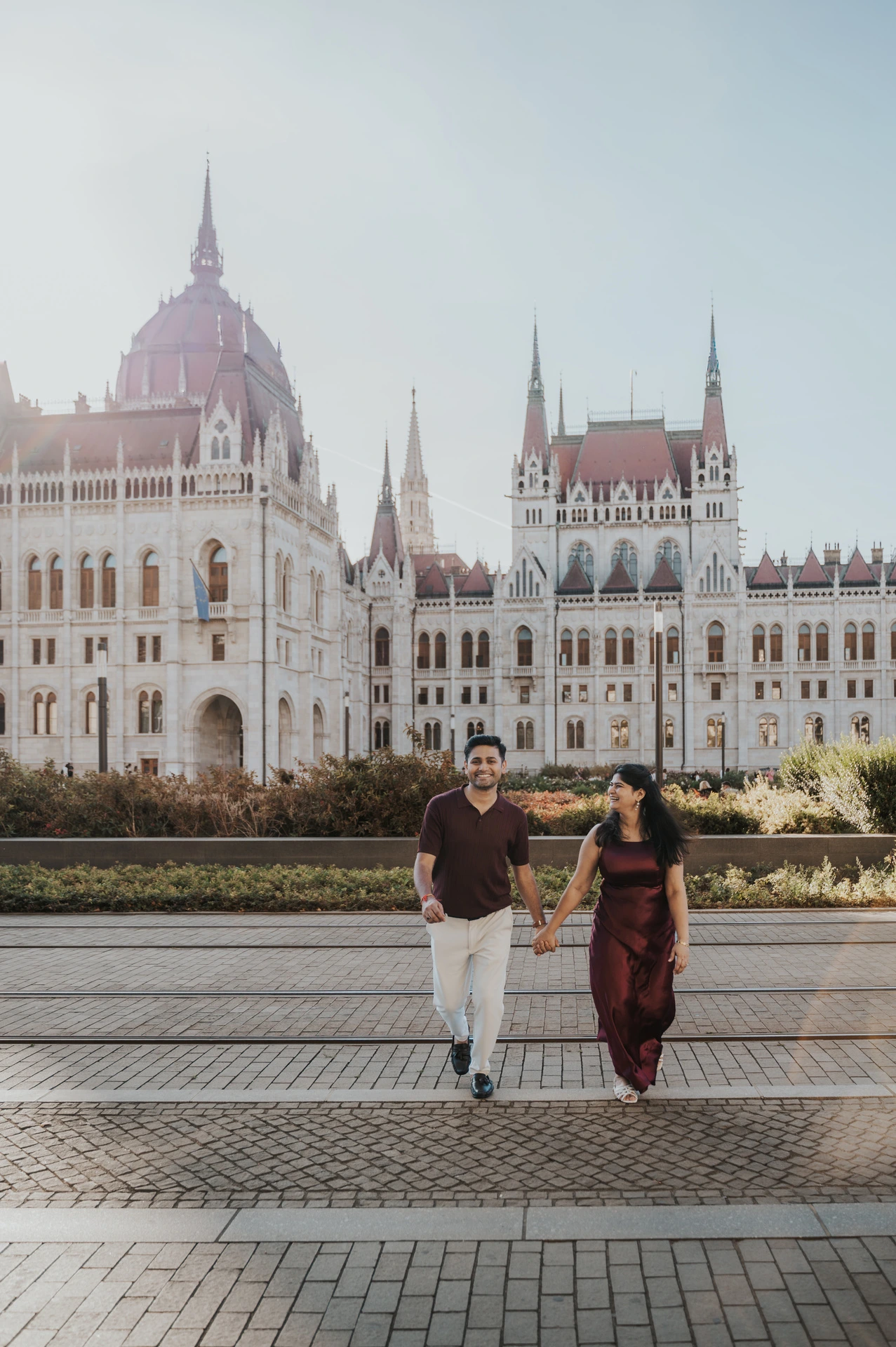 The couple walking hand in hand along the paving beside Parliament’s flowerbeds during their couple photoshoot in Budapest.